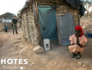 richard hotes surveying village while older villager squats in front of the mud hut
